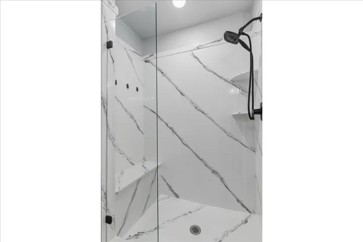 Walk-in shower featuring glass enclosure, white walls with black veining, two built-in corner shelves, and matte black fixtures