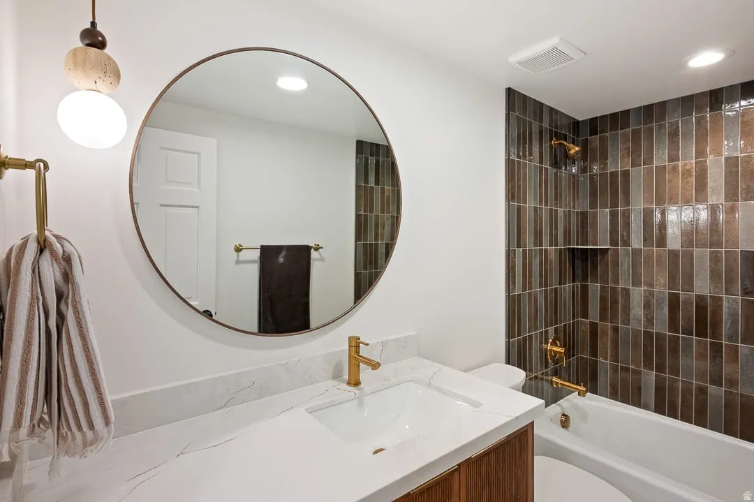 Bathroom featuring a circular mirror, a white rectangular sink, a gold-finish faucet, a white countertop, and vertical brown and gray shower tiles