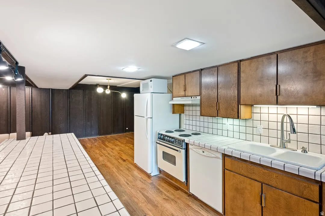 Kitchen area featuring wood-finish cabinetry, white tile countertops and backsplash, a double basin sink with a high-arc faucet, and white appliances including a refrigerator and electric range