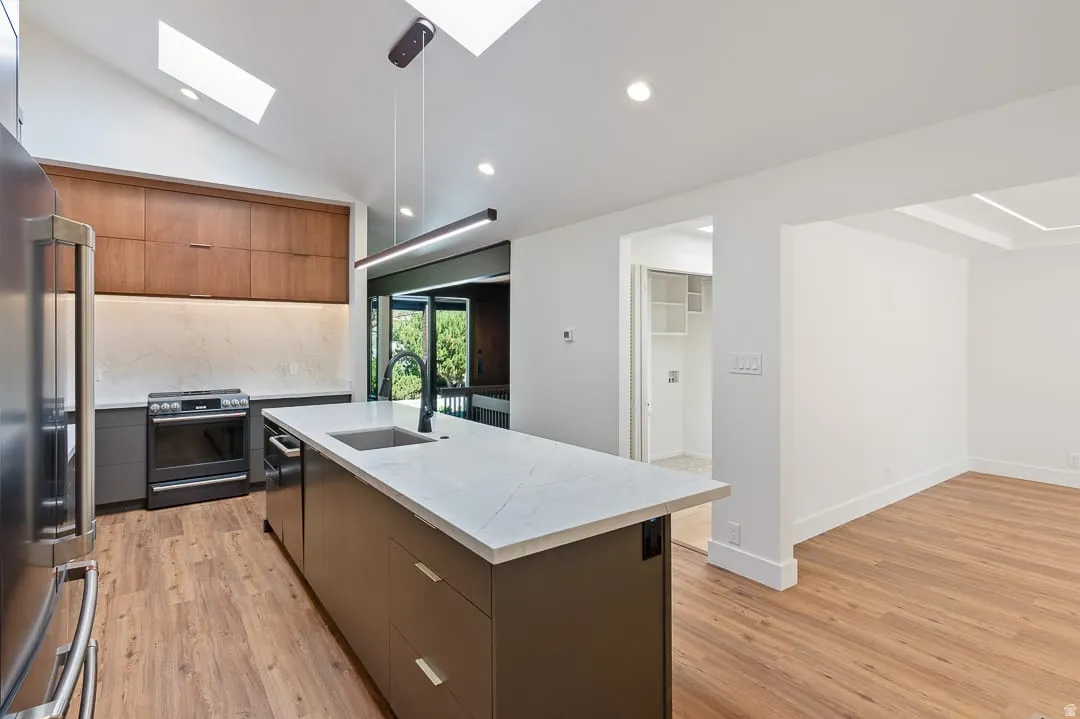 Modern kitchen featuring a vaulted ceiling with skylights, wood-finish flooring, a central island with an integrated sink, stainless steel appliances, and a sophisticated two-tone cabinetry design