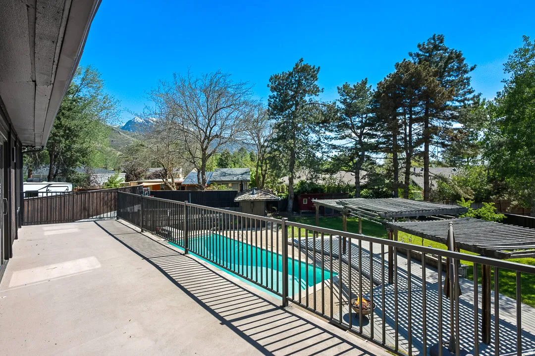 Outdoor deck with dark metal railing overlooking a fenced swimming pool, two wood pergolas, and mature trees