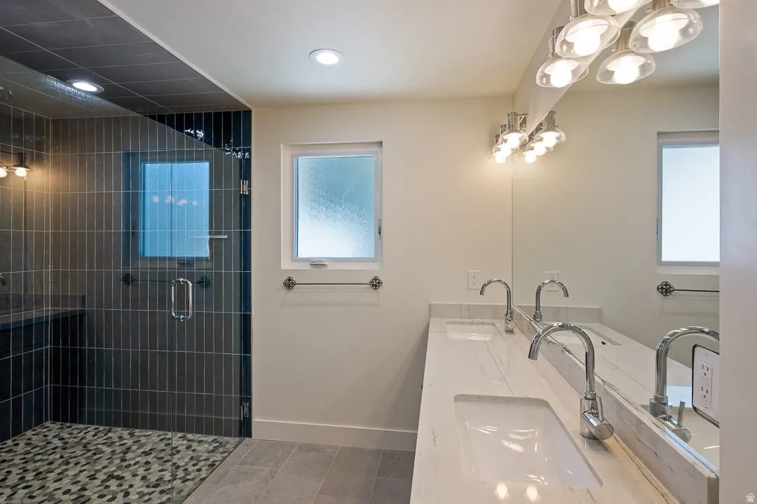 Bathroom featuring a dual vanity with integrated sinks and stone-finish countertops, an oversized mirror, brushed nickel light fixtures, and a spacious glass-enclosed shower with vertical tile detailing and a stone pebble floor