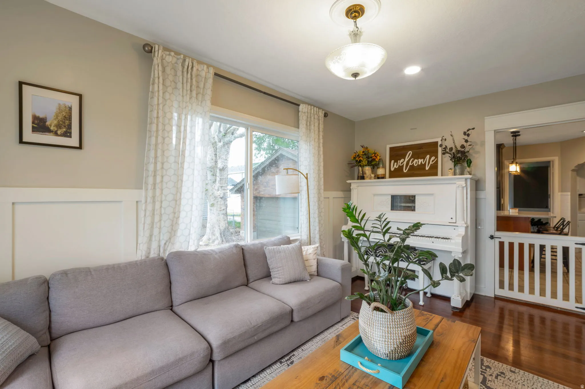 Bright living space featuring warm wood-finish flooring, wainscoting, and large windows with privacy drapes