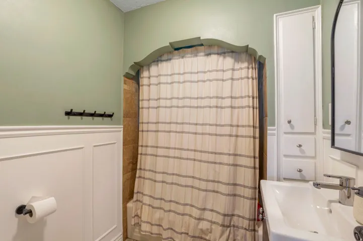 Bathroom featuring wainscoting, an arched shower surround with tile, a pedestal sink with a modern faucet, and built-in cabinetry