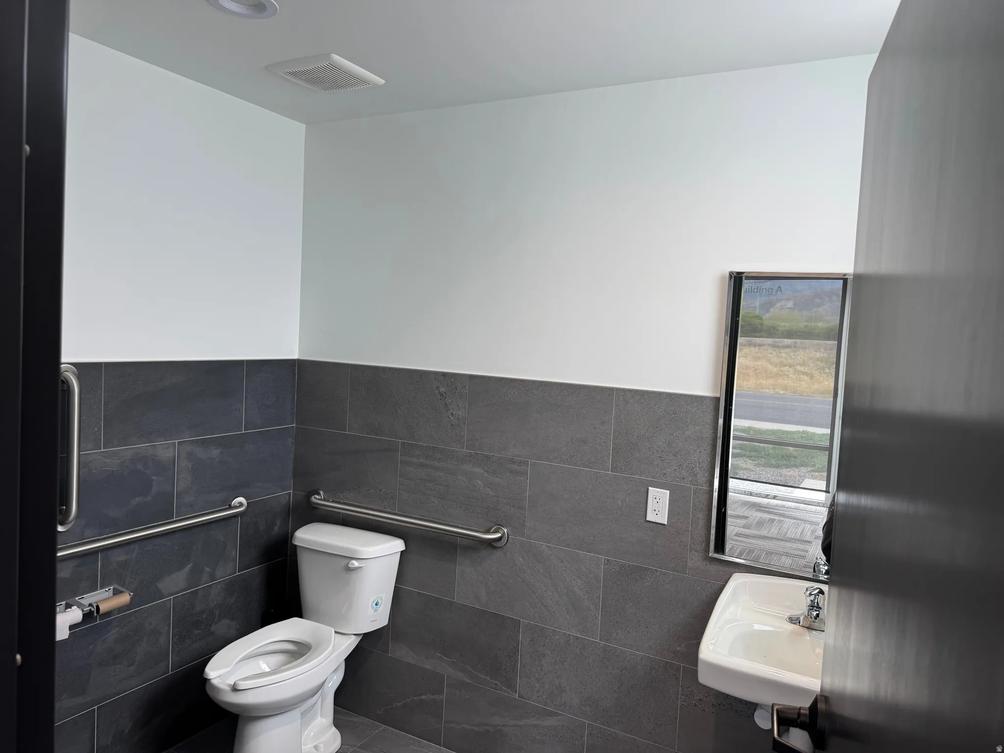 Bathroom featuring dark gray tile walls and flooring, white toilet, white wall-mounted sink, and stainless steel grab bars