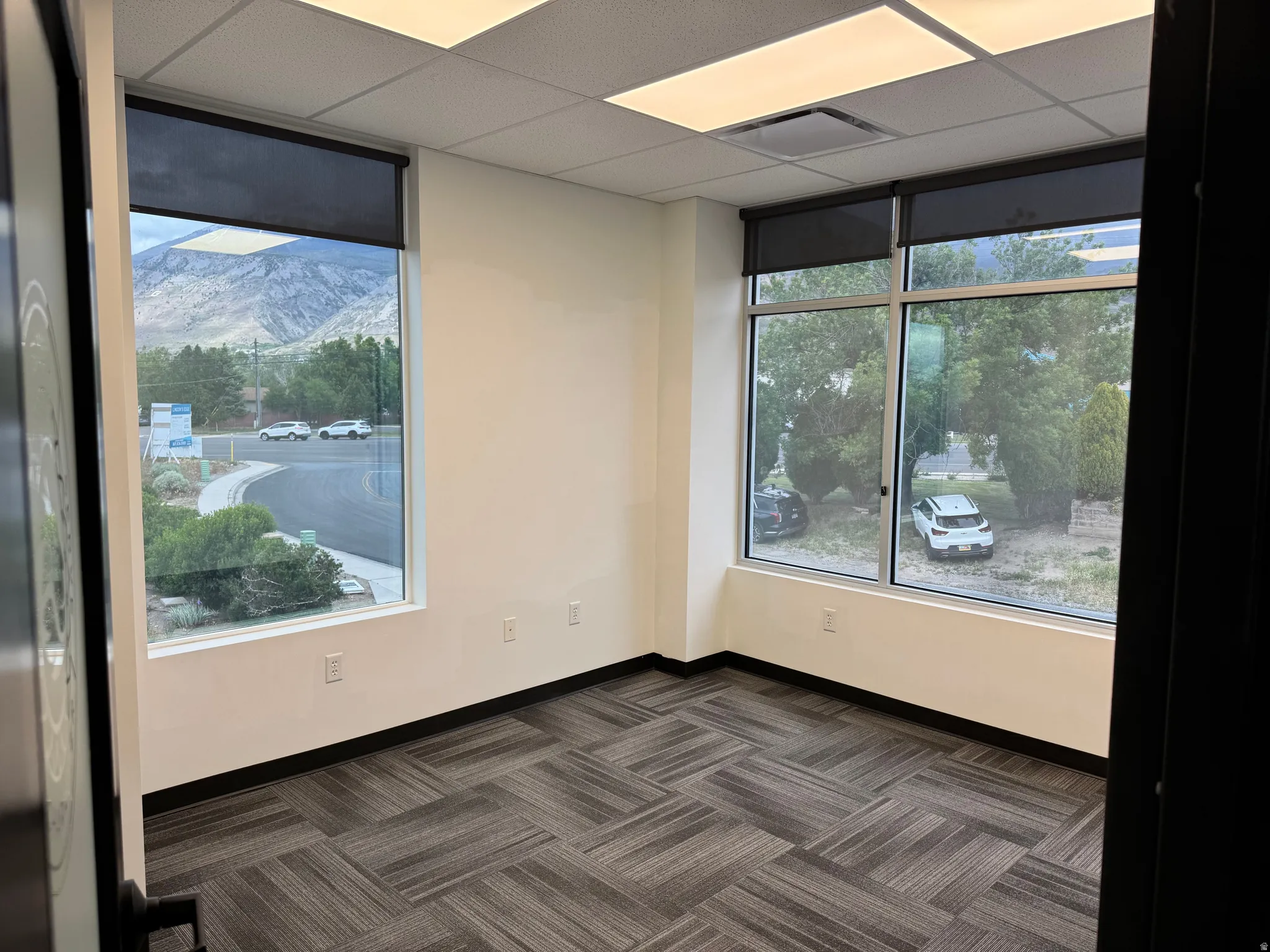 Interior office space featuring large windows with mountain scenery, patterned carpet flooring, recessed ceiling lighting, roller shades, and multiple electrical outlets