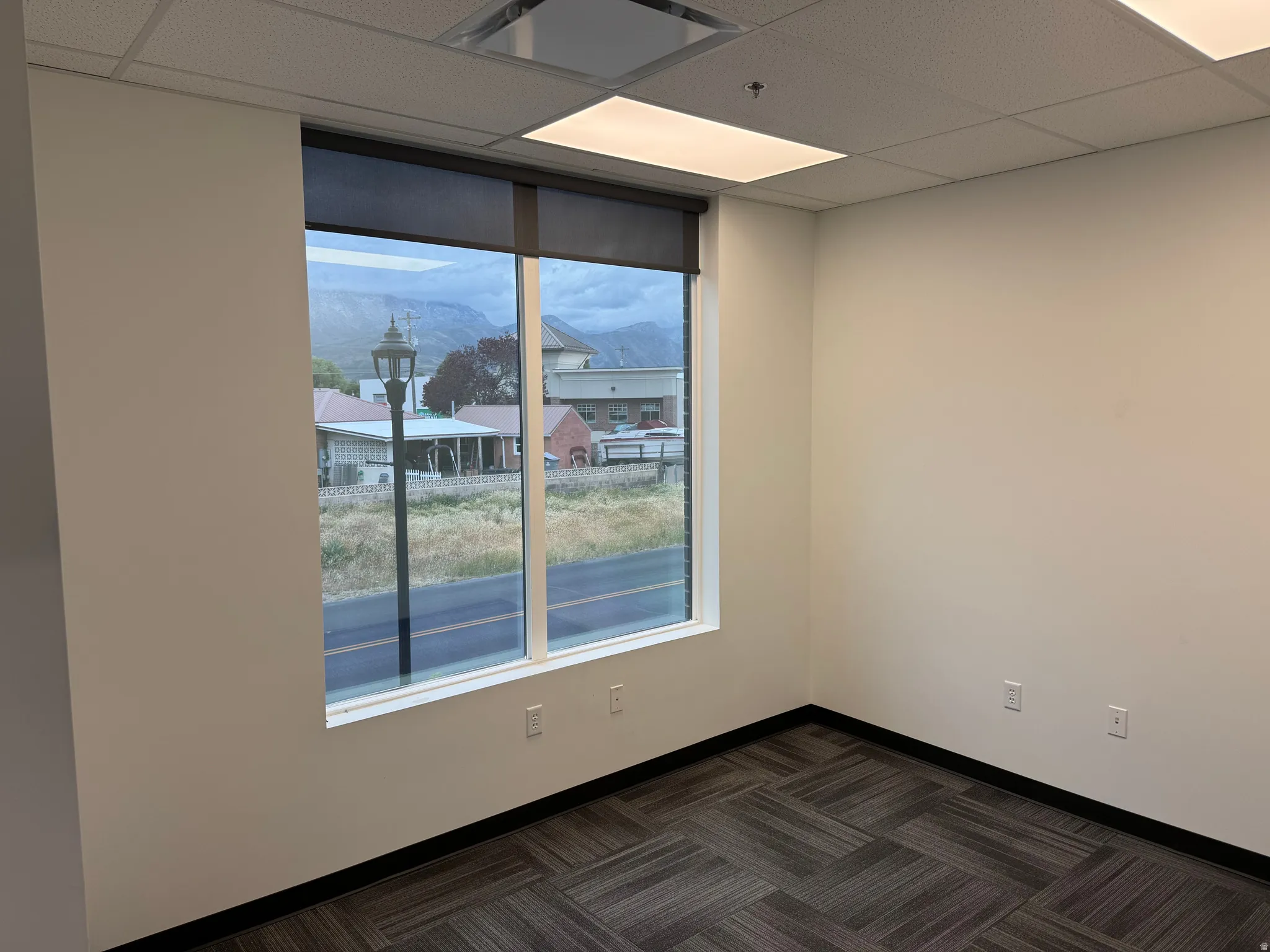 Bright interior space featuring a large window with roller shades, neutral tone walls, patterned carpet tile flooring, and a drop ceiling with integrated lighting