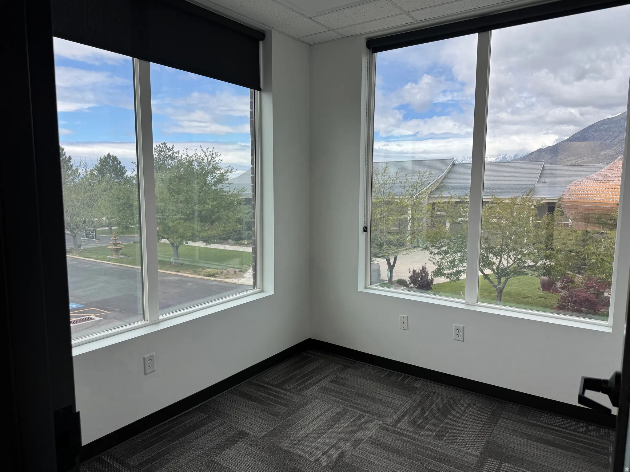 Corner room featuring two large windows, roller shades, white walls, and geometric pattern carpet flooring