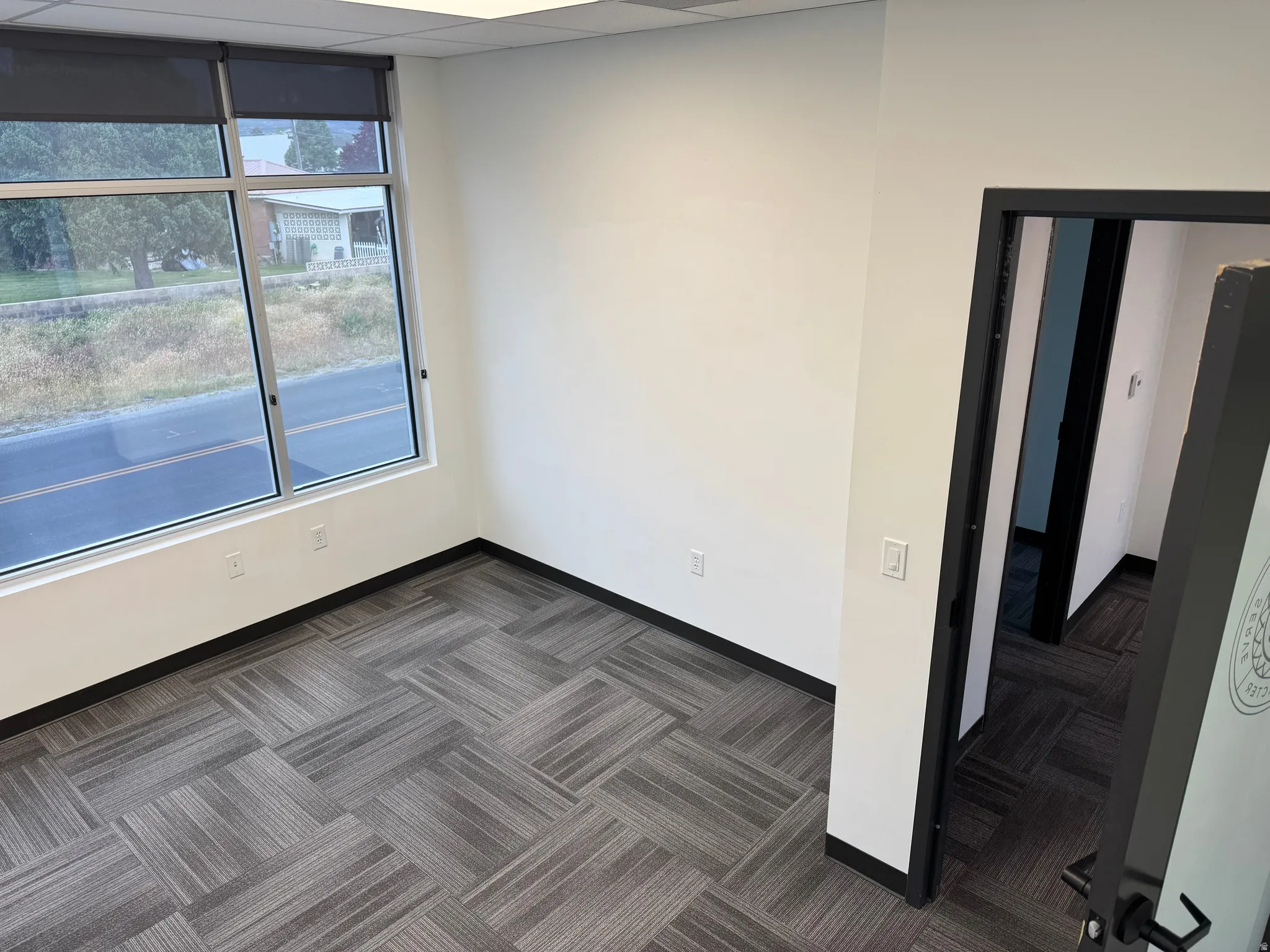 Bright interior space featuring a large window with roller shades, neutral wall finishes, patterned carpet tile flooring, recessed lighting, and a dark-framed doorway
