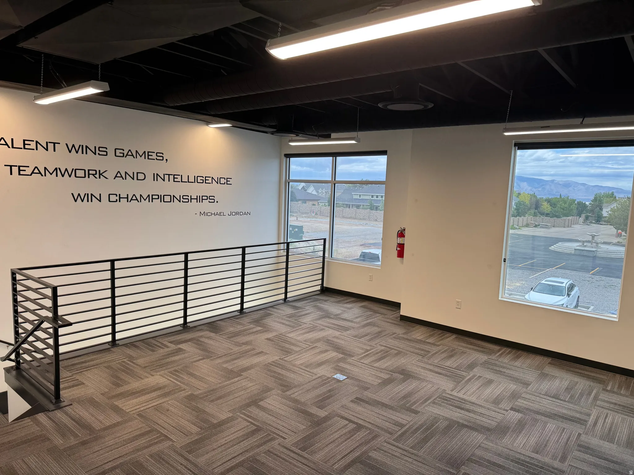Spacious open-concept commercial space featuring large windows, modern track lighting, durable patterned carpet tile flooring, and a contemporary black metal stair railing