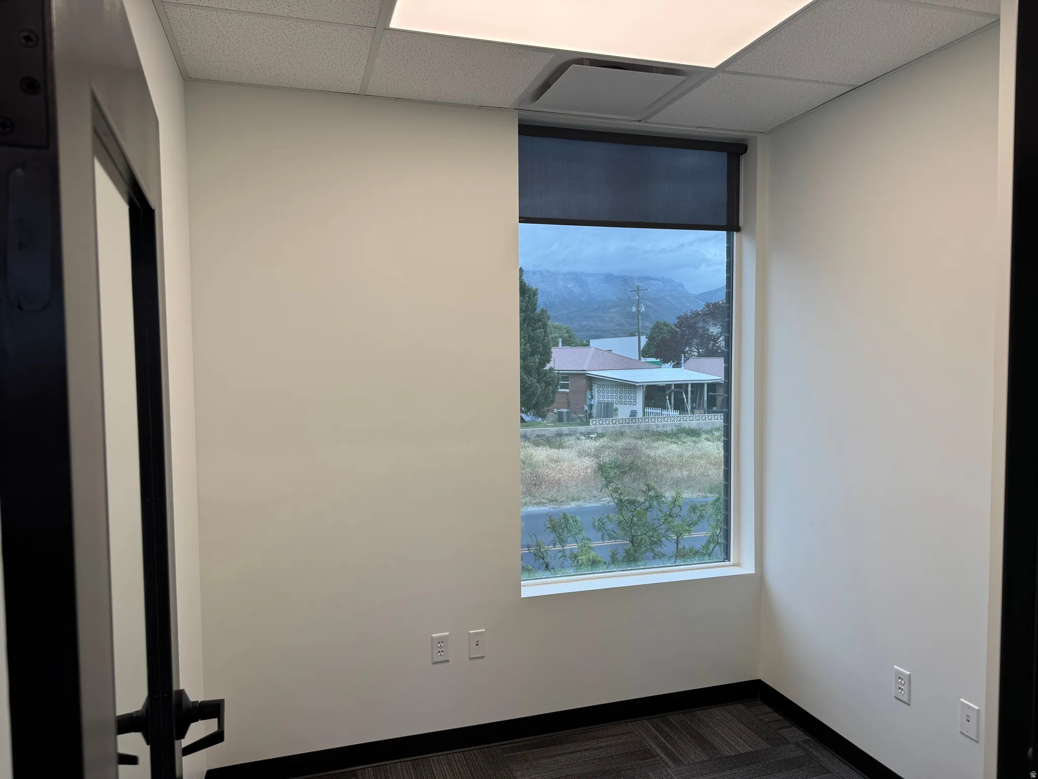 Bright interior space featuring a window with roller shade, white walls, and geometric pattern carpet flooring