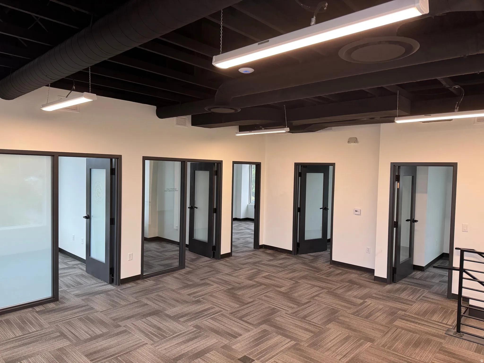 Open-concept office space featuring a black exposed ceiling with ductwork, multiple private offices with frosted glass doors, linear overhead lighting, and patterned carpet flooring