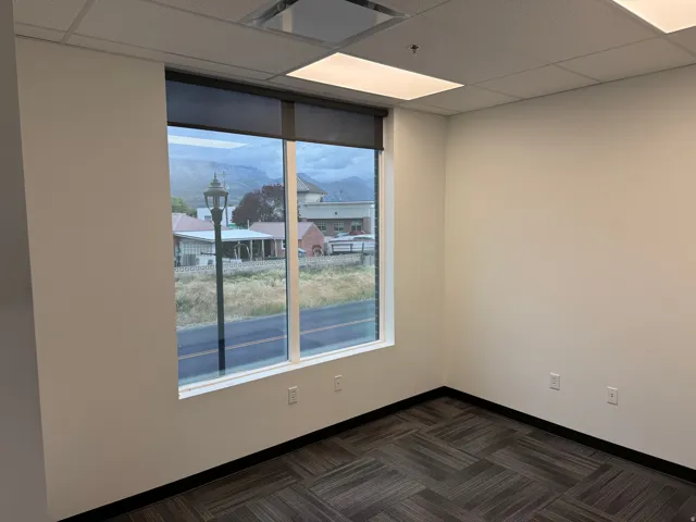 Bright interior space featuring a large window with roller shades, neutral tone walls, patterned carpet tile flooring, and a drop ceiling with integrated lighting