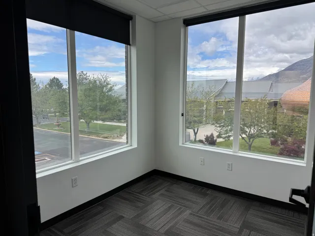 Corner room featuring two large windows, roller shades, white walls, and geometric pattern carpet flooring
