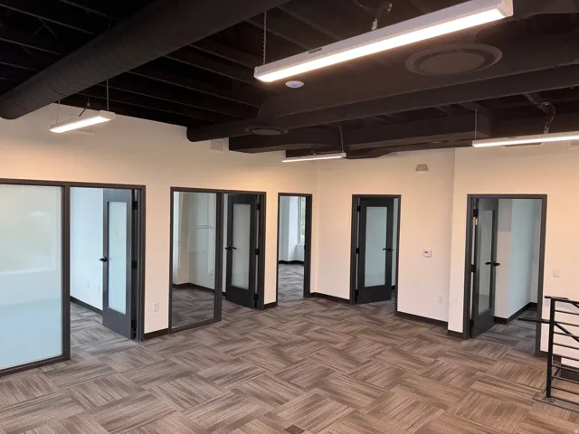 Open-concept office space featuring a black exposed ceiling with ductwork, multiple private offices with frosted glass doors, linear overhead lighting, and patterned carpet flooring