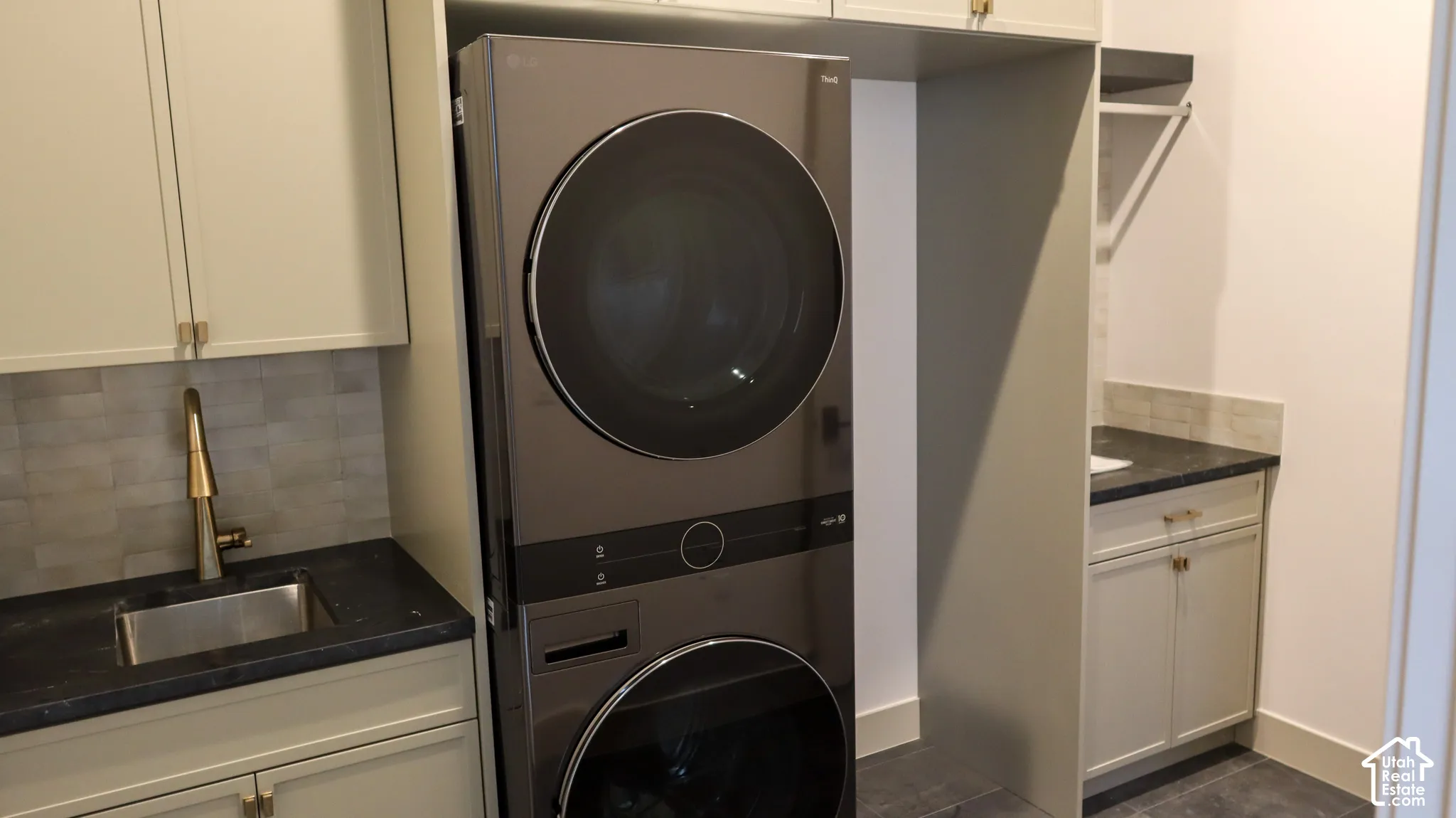 Integrated stacked washer and dryer, under-mount stainless steel sink, dark stone-finish countertop, light neutral cabinetry, and subway tile backsplash