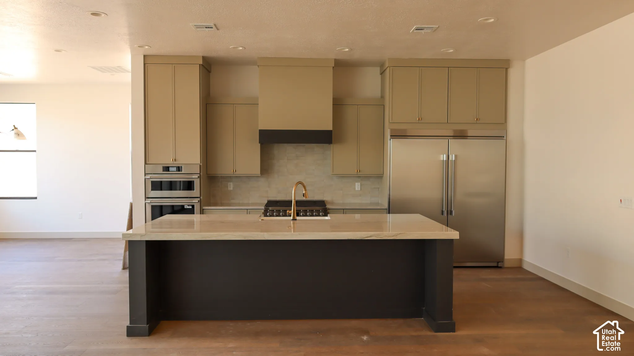 Kitchen featuring an expansive island with a light-toned countertop, stainless steel double ovens, a built-in refrigerator, a gas range with a range hood, and wood-finish flooring