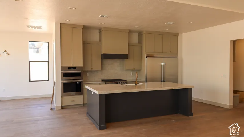 Open-concept kitchen featuring an island with a light-colored countertop, stainless steel appliances, built-in double ovens, and wood-finish flooring