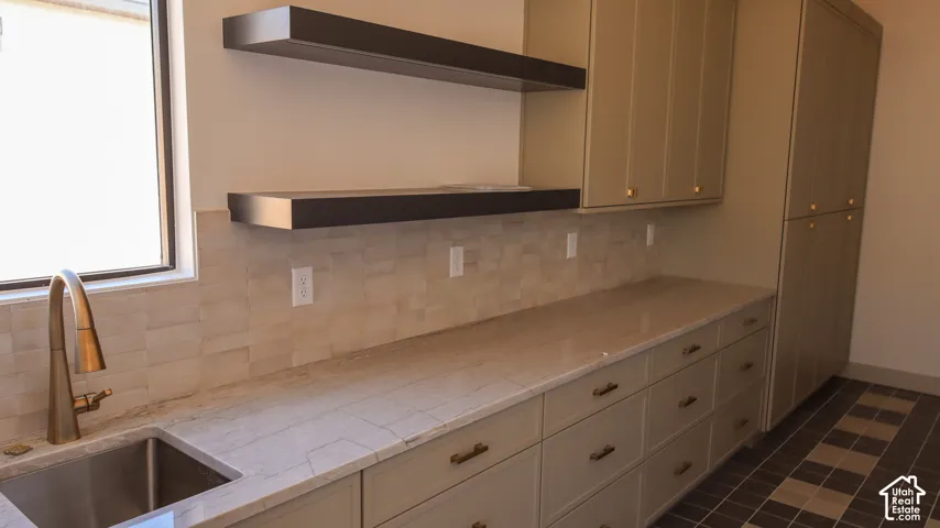 Built-in cabinetry with brass hardware, undermount sink with gooseneck faucet, light-toned backsplash, floating shelves, and window