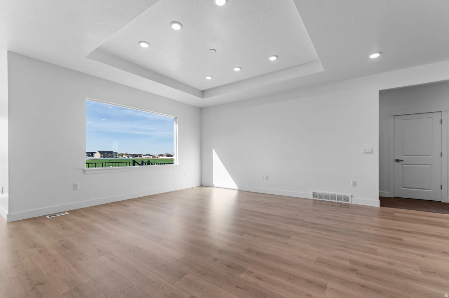 Spacious room featuring wood-finish flooring, a tray ceiling with recessed lighting, and a large window overlooking distant properties
