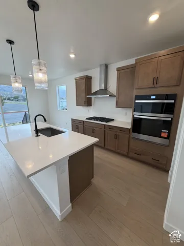 Kitchen featuring wood-finish cabinetry, quartz countertops, a central island with an undermount sink, built-in double wall ovens, and a stainless steel range hood