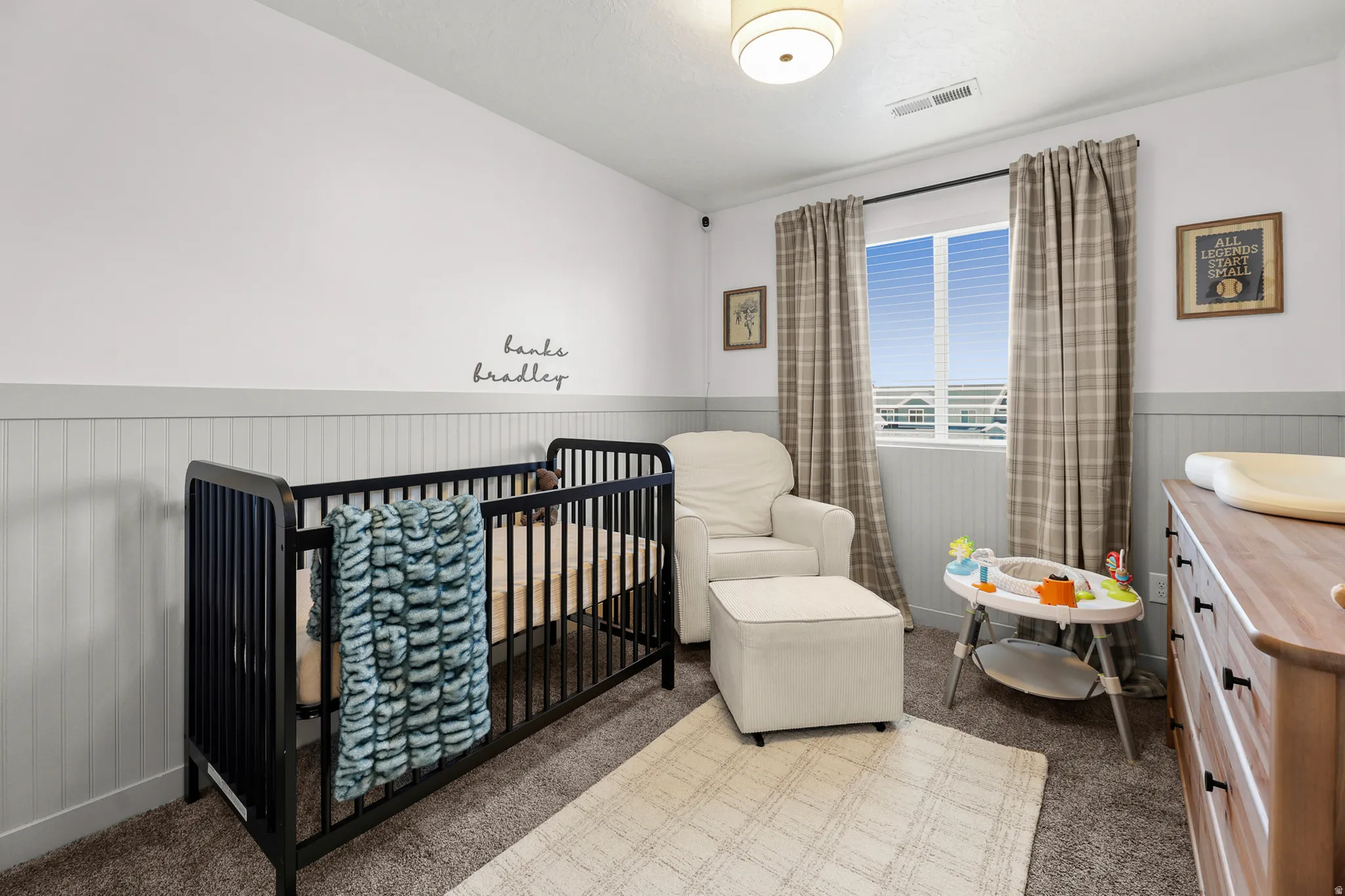 Room featuring white and gray wainscoting, neutral carpet, a single window with blinds and plaid curtains, and a flush mount ceiling light