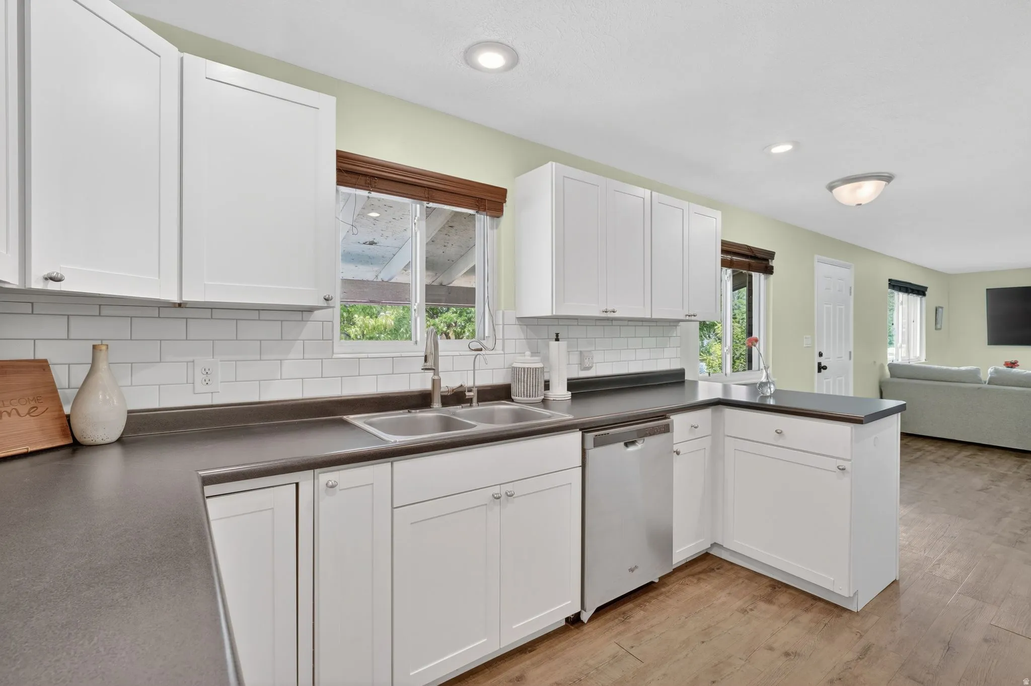 Kitchen featuring white shaker cabinetry, dark countertops, a stainless steel double basin sink, subway tile backsplash, and wood-finish flooring