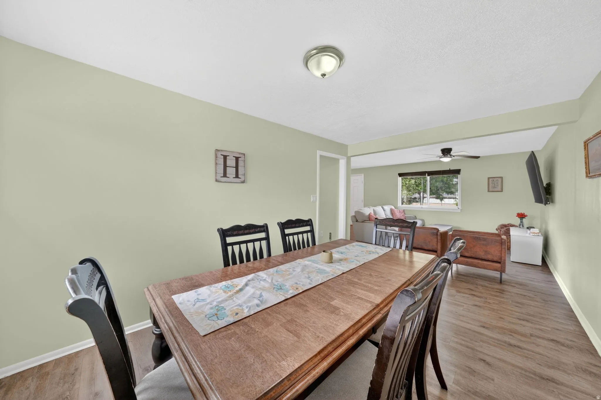 Dining area featuring light green walls, wood-finish flooring, a ceiling-mounted light fixture, and an open layout providing sightlines into an adjacent living space
