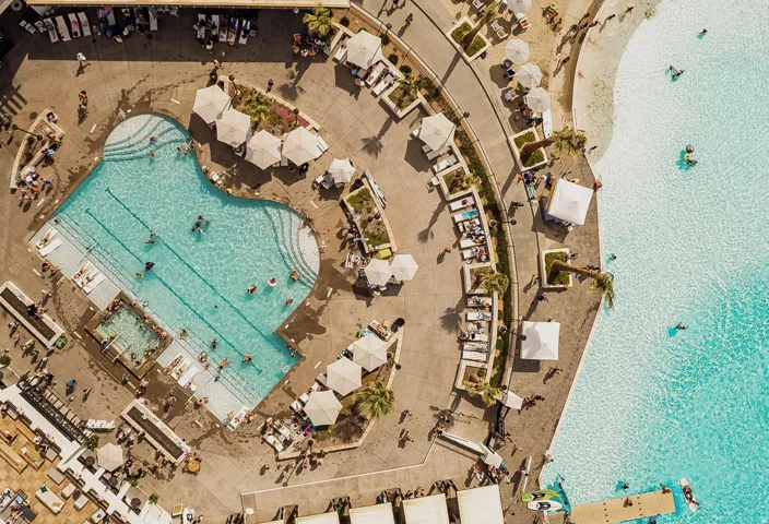Expansive aquatic recreation area featuring a large resort-style pool, a sand-lined lagoon with clear blue water, sunbathing decks, multiple shade umbrellas, and mature palm trees
