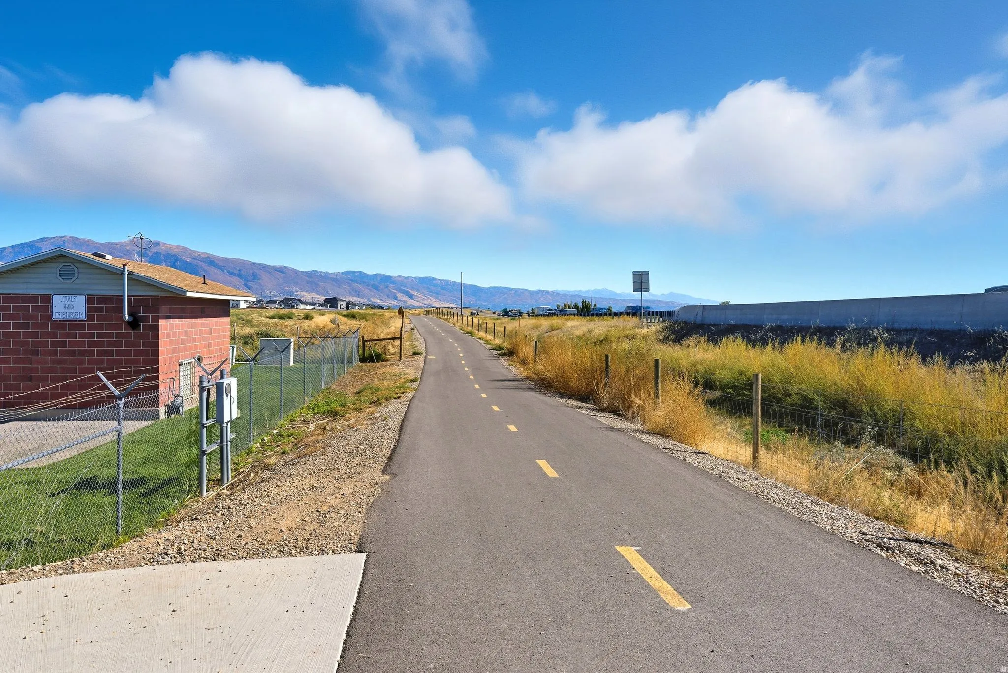 Paved pathway bordered by natural landscaping, featuring distant mountains and a clear sky backdrop