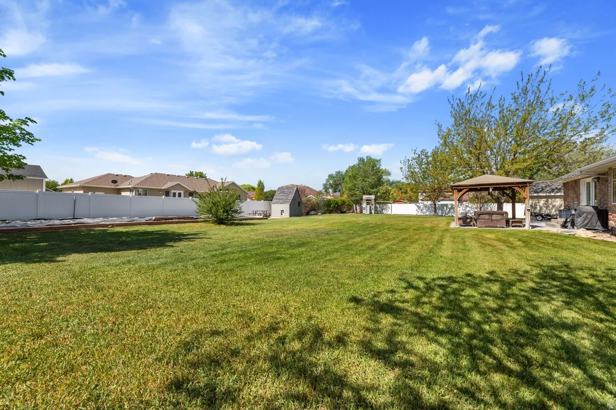 Expansive backyard featuring a large green lawn, a white privacy fence, a wooden gazebo, a storage shed, and mature trees