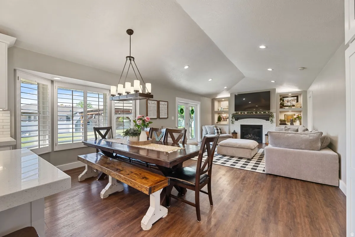 Open-concept living space featuring wood-finish flooring, a contemporary fireplace with built-in shelving, vaulted ceilings, and large bay windows with plantation shutters