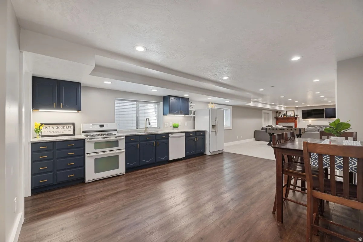 Spacious open-concept lower level featuring rich wood-finish flooring, a full kitchen with dark cabinetry and light countertops, a white double oven range, white refrigerator, and recessed lighting