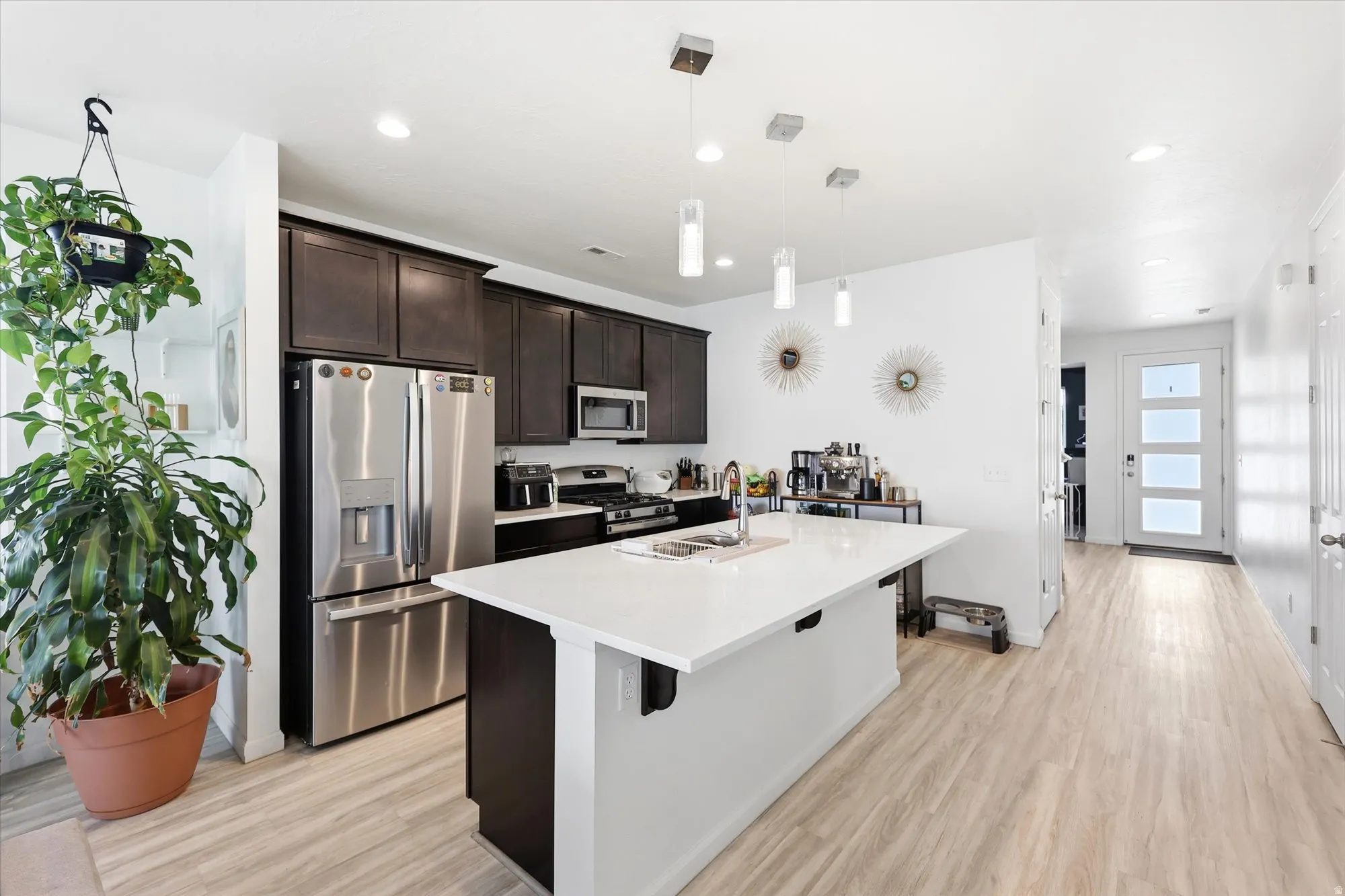 Contemporary kitchen featuring an expansive island with a white countertop, dark wood-finish cabinetry, stainless steel appliances, pendant lighting, and light wood-finish flooring