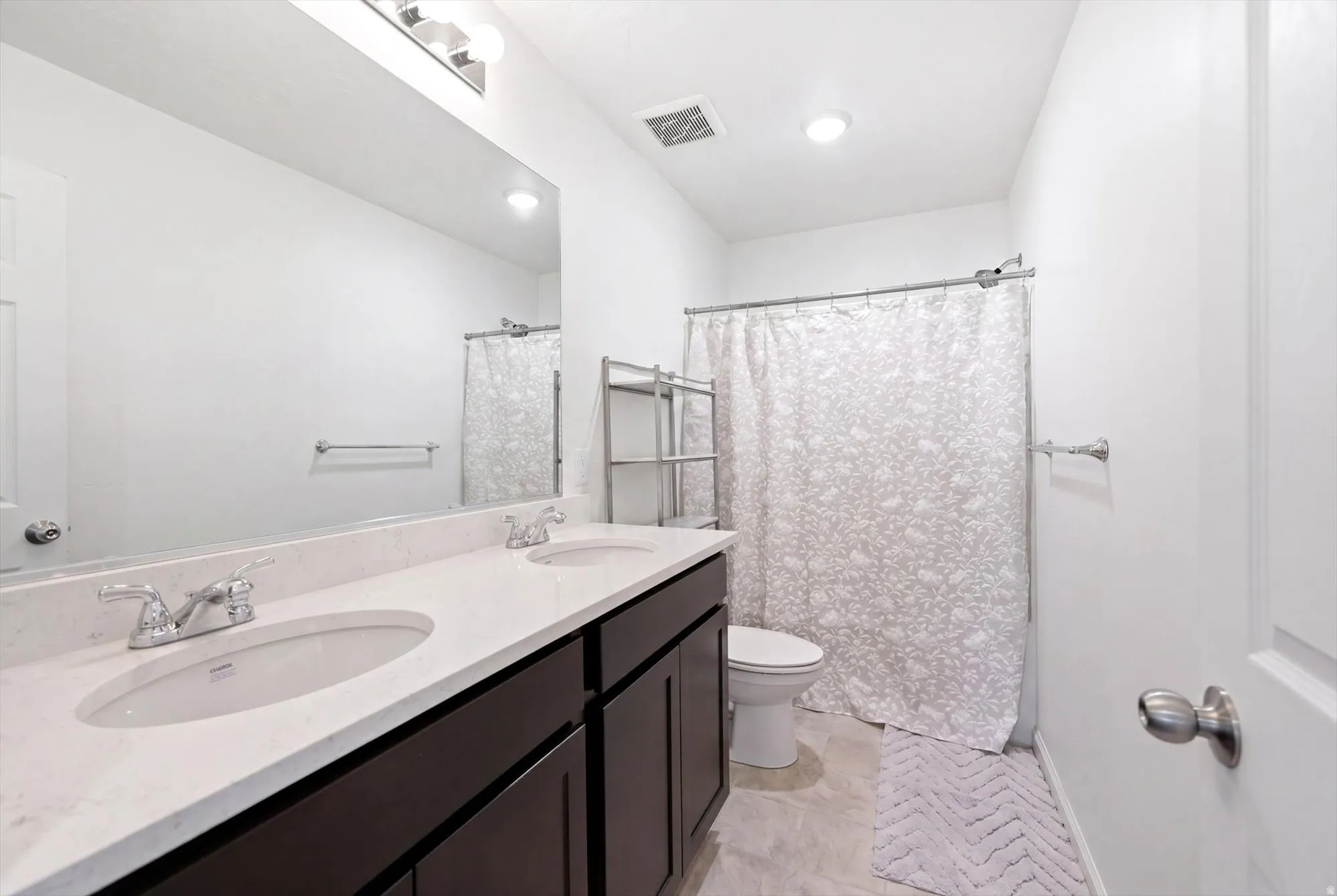 Double vanity with integrated sinks and chrome fixtures, dark wood-finish cabinetry, a full-width mirror, recessed ceiling lighting, and a shower-tub combination
