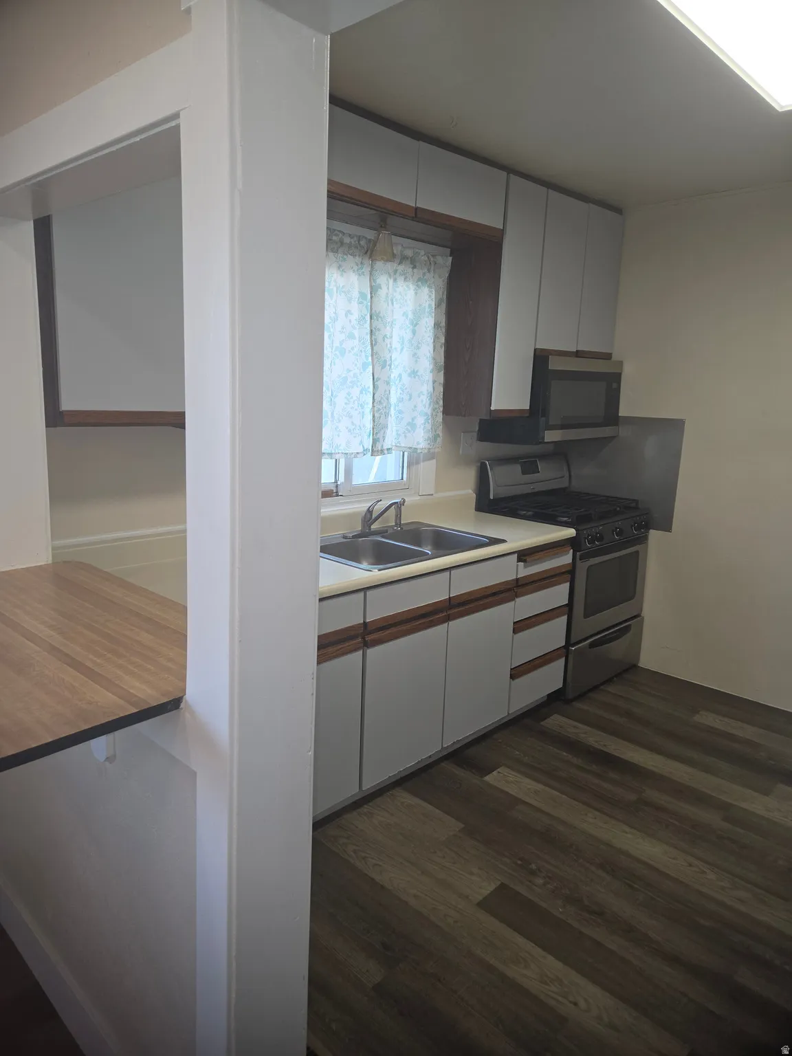 Kitchen featuring wood-finish flooring, white cabinetry with wood accents, double basin stainless steel sink, stainless steel gas range, and a built-in microwave