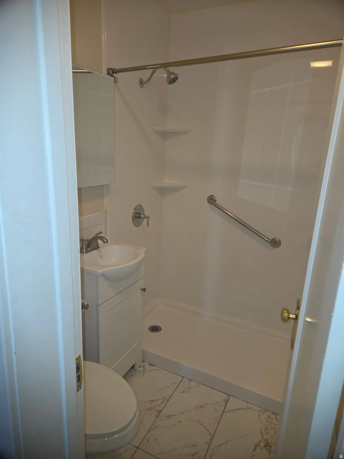 Compact bathroom featuring a white shower stall with built-in shelving and a grab bar, a white pedestal sink with brushed nickel faucet, a white toilet, and marble-look tile flooring
