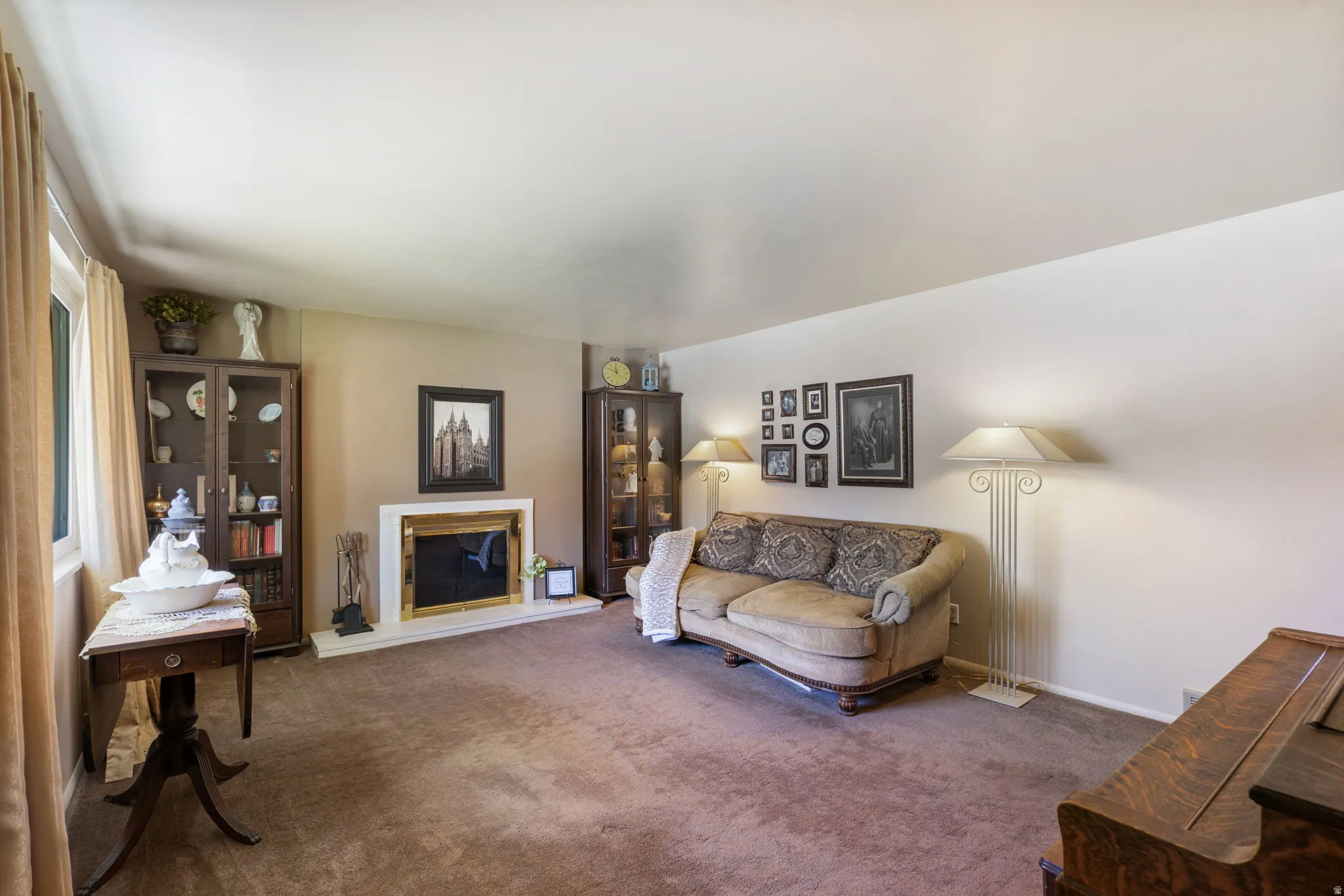 Carpeted living area featuring a fireplace with a white surround and brass-toned insert, a window with drapery, and two built-in display cabinets