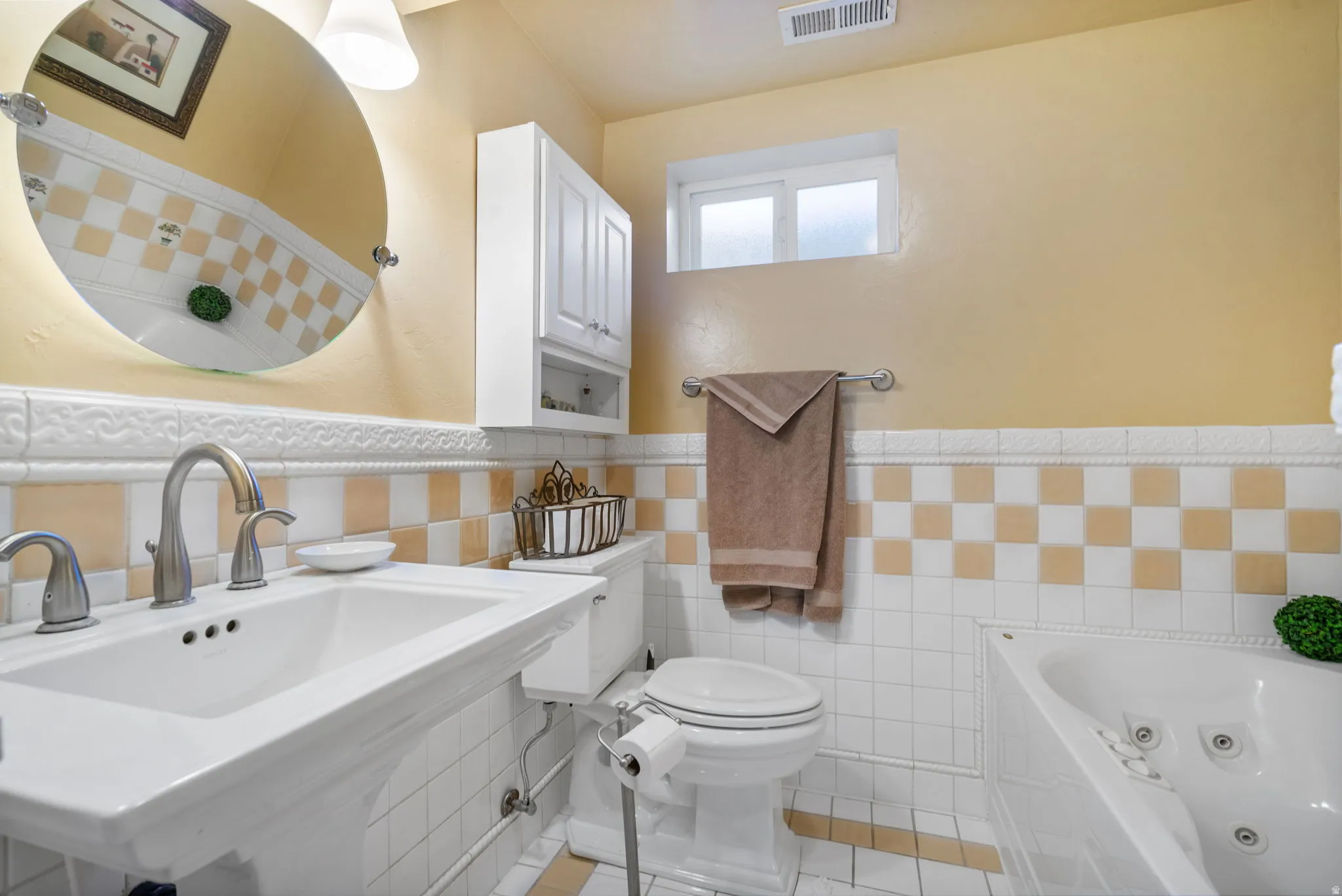 Bathroom featuring a pedestal sink with dual-handle faucet, a jetted soaking tub, white tiled walls with a checkerboard accent, a white toilet, and a medicine cabinet