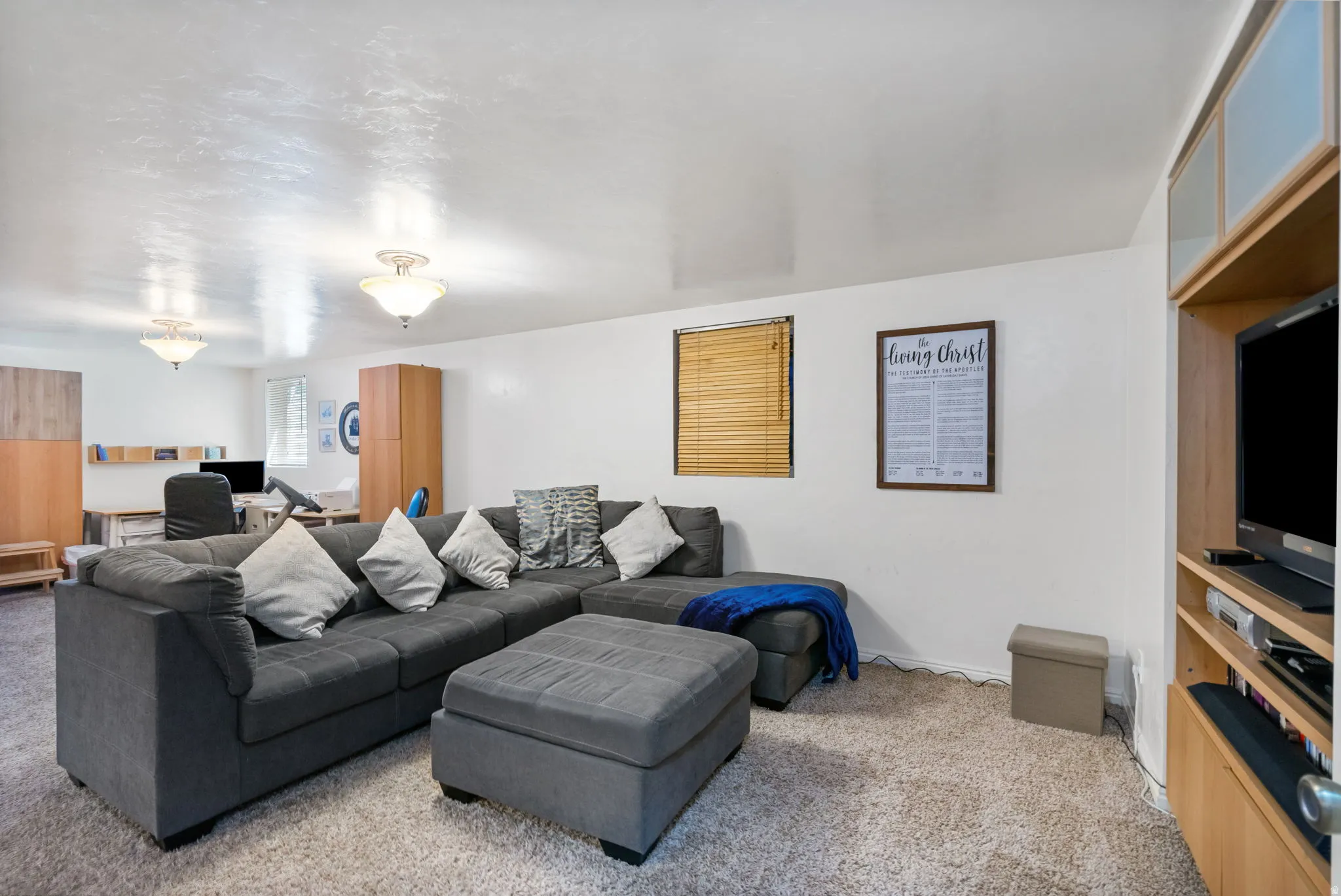 Carpeted room featuring a built-in entertainment center, two ceiling-mounted light fixtures, a window with horizontal blinds, and a built-in desk area with shelving