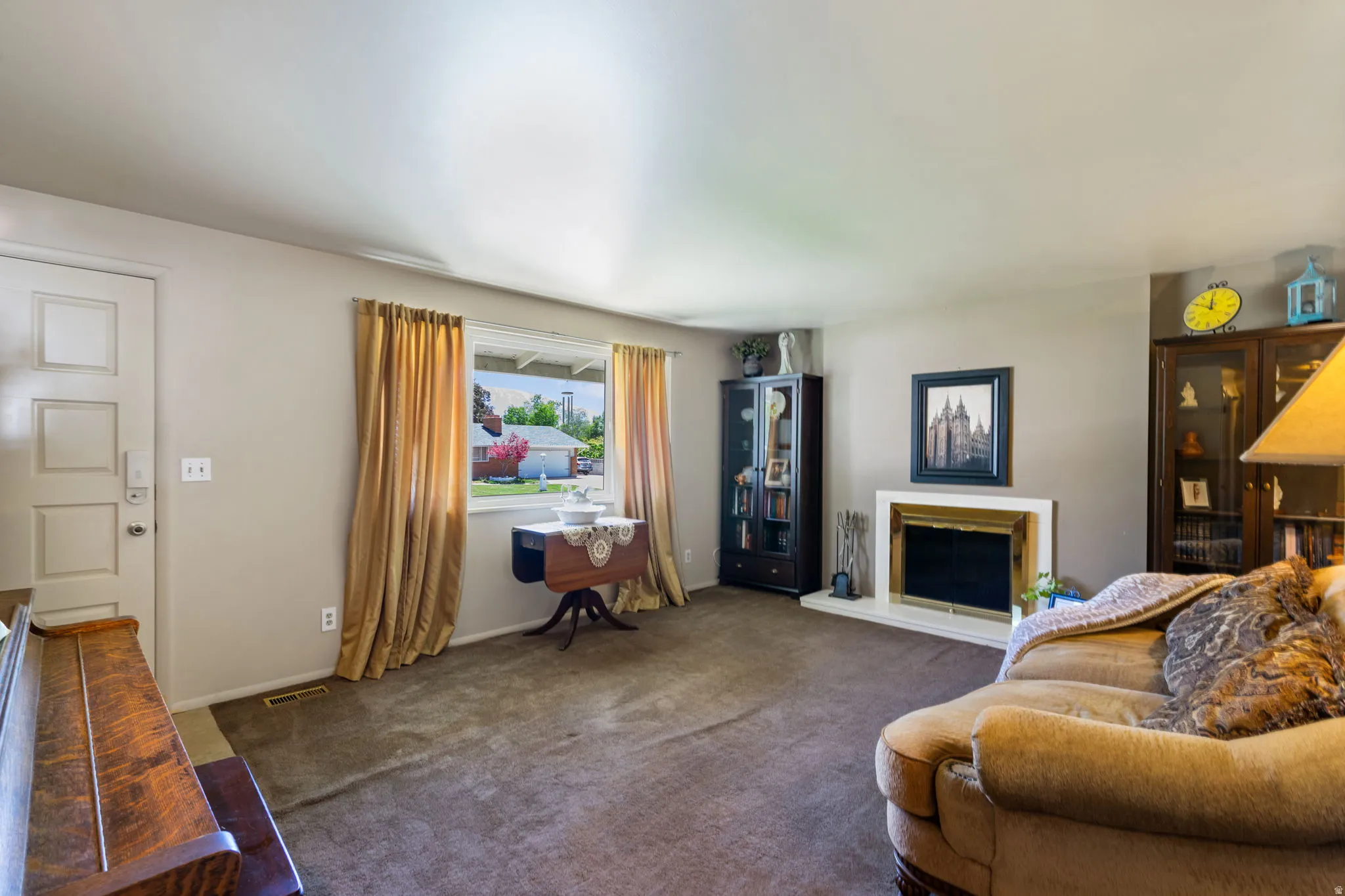Spacious living area featuring a fireplace with a white mantel, large window with gold drapes, light tan wall color, and brown carpeting