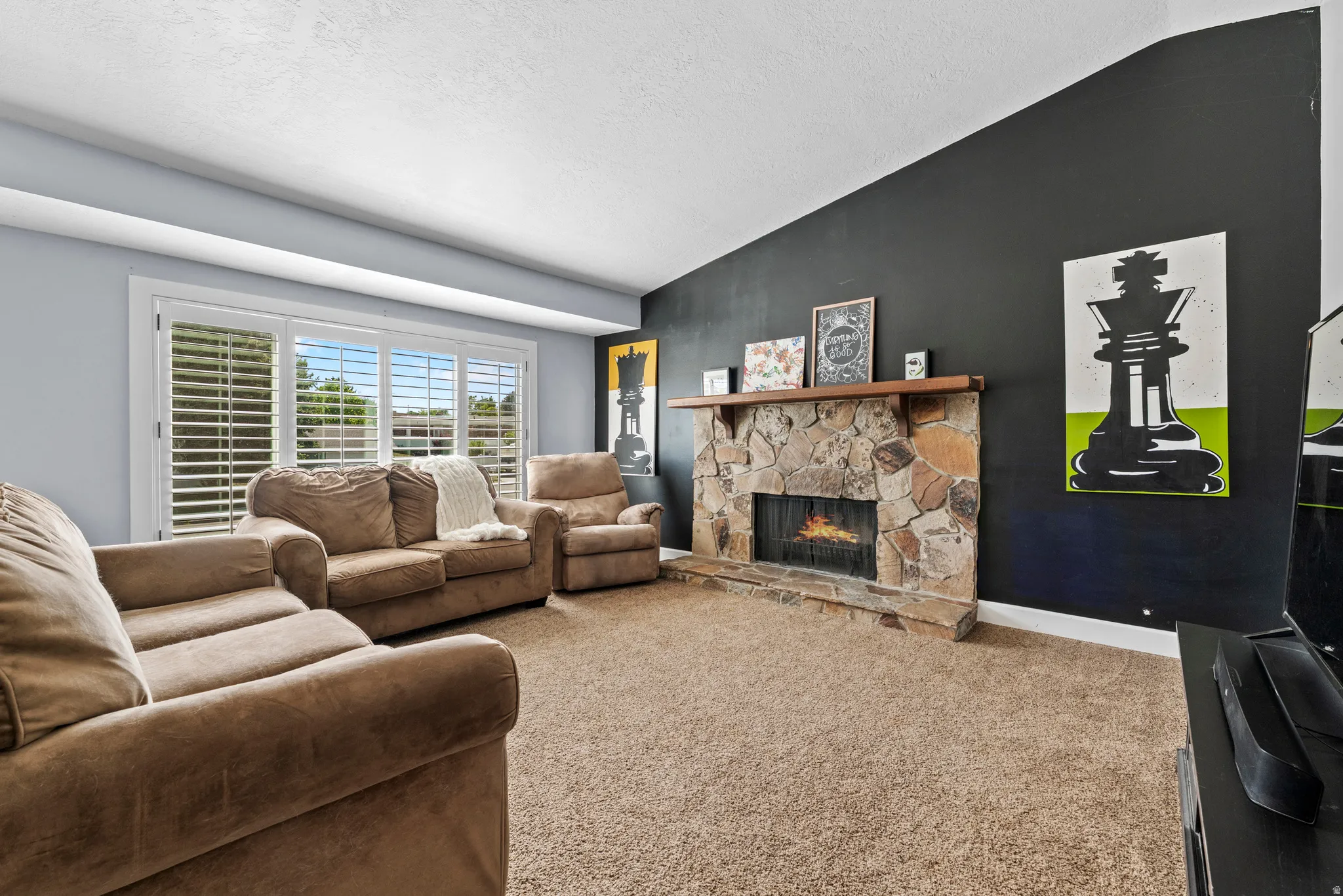 Stone fireplace with a wood mantle, carpet flooring, large windows with white louvered shutters, and light blue and dark accent walls