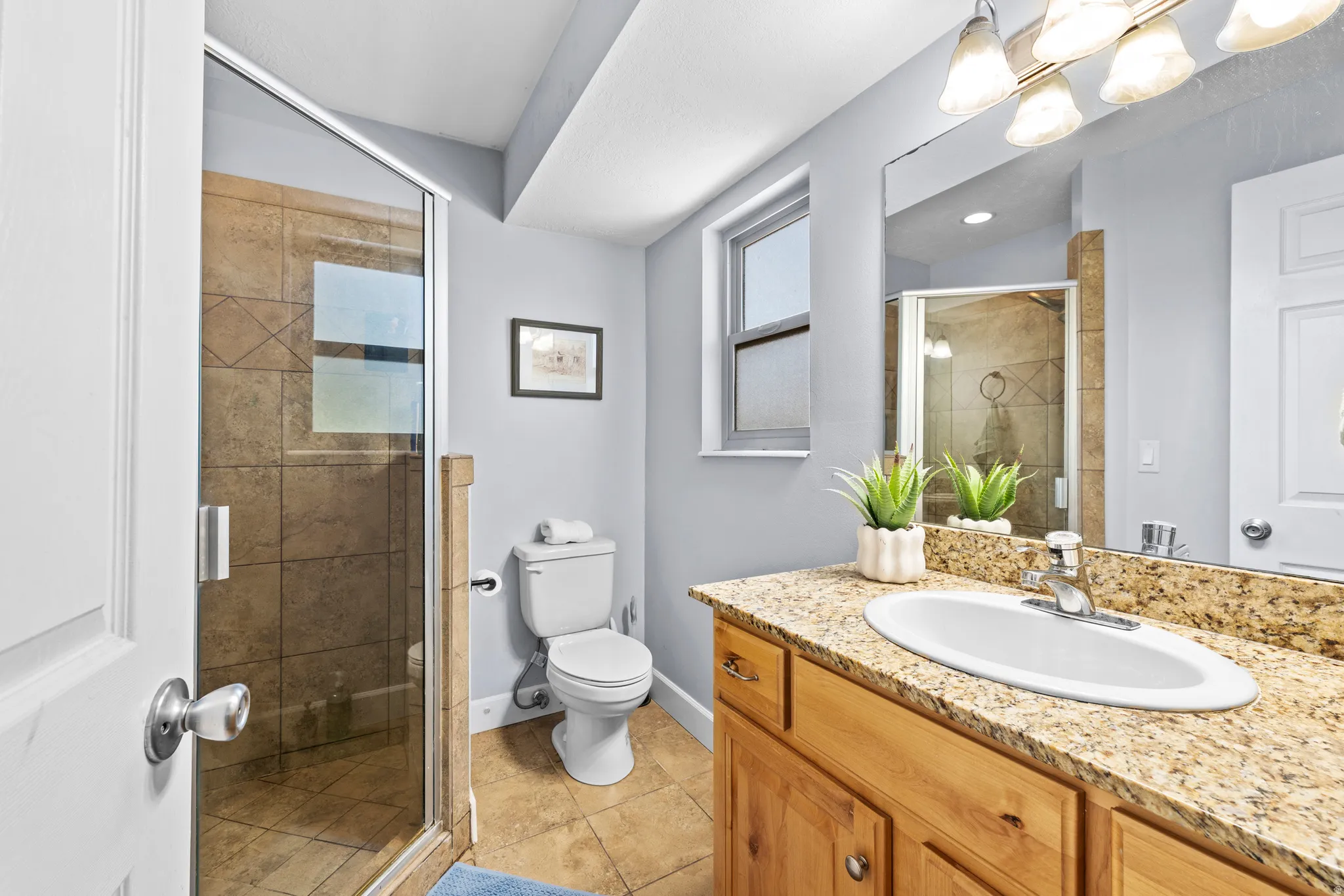 Full bathroom featuring a glass-enclosed shower with tile surround, a single vanity with a light-colored stone countertop, wood-finish cabinetry, and tiled flooring