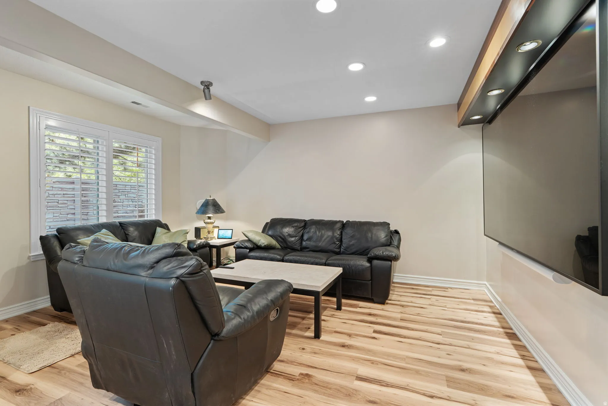 Spacious lounge with wall television featuring wood-finish flooring, recessed ceiling lighting, and a window with plantation shutters
