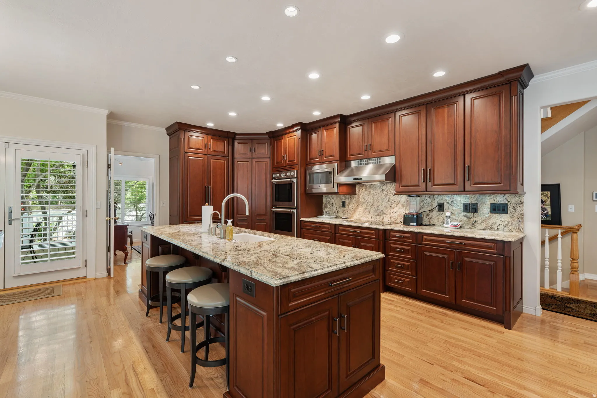 Gourmet kitchen featuring a large central island with seating, extensive mahogany wood cabinetry, light-toned granite countertops with a matching backsplash, stainless steel appliances, Thermador double ovens and integrated refrigerator and dishwasher