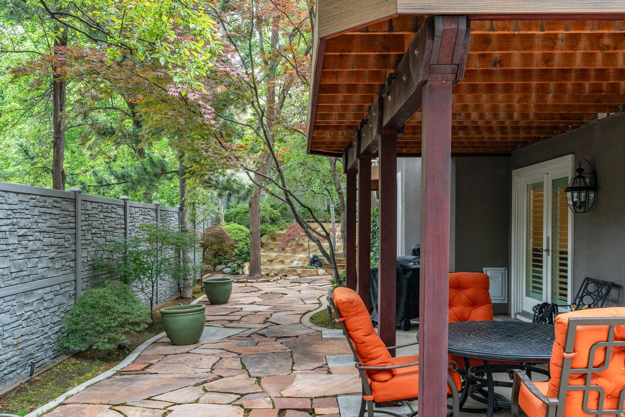 Expansive custom cut stone patio with a covered outdoor living area featuring a wood-beam ceiling, decorative stone-look privacy fence, and mature landscaping