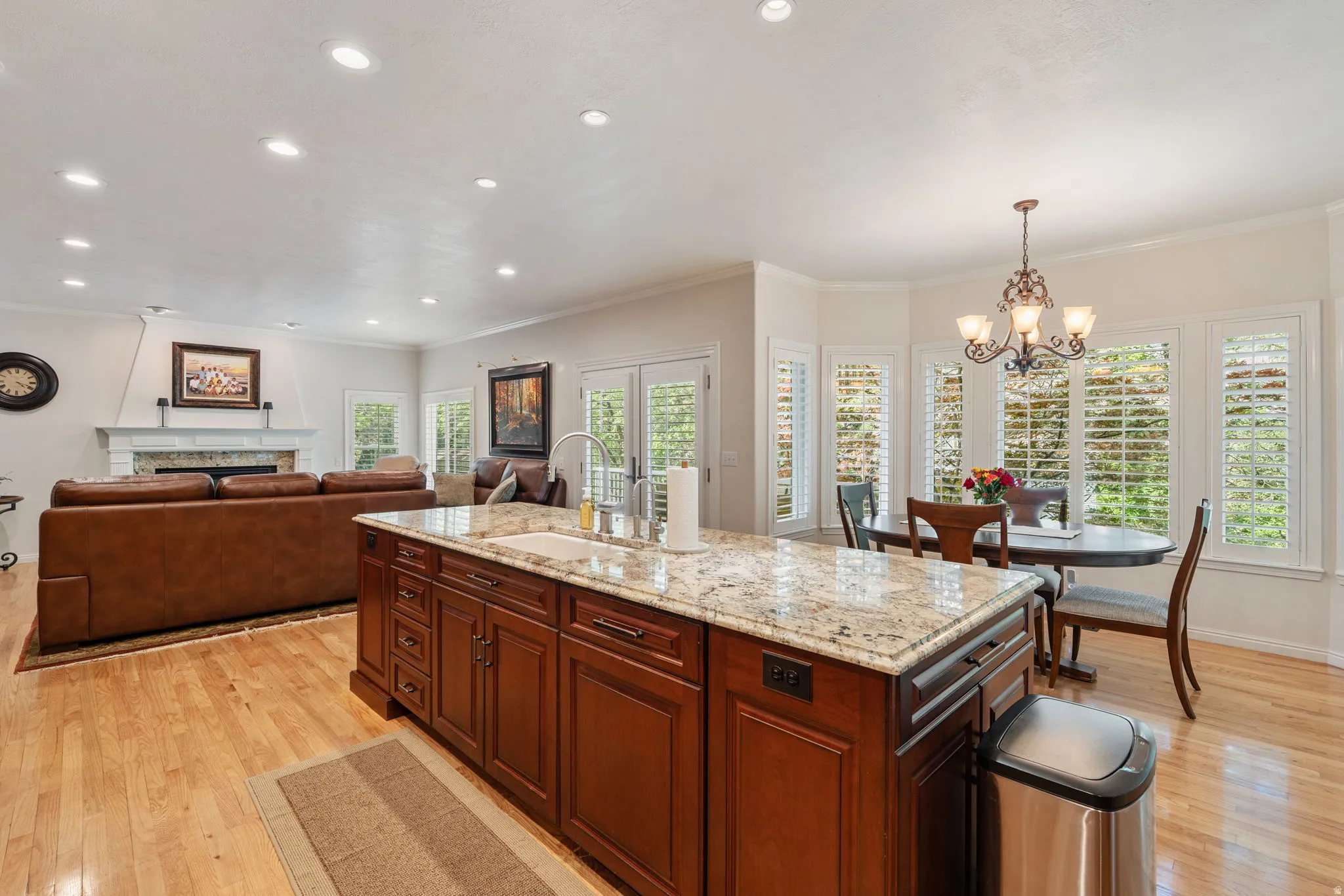 Kitchen island with granite countertop and integrated sink, hardwood flooring, recessed lighting, and a breakfast area with multiple windows featuring plantation shutters