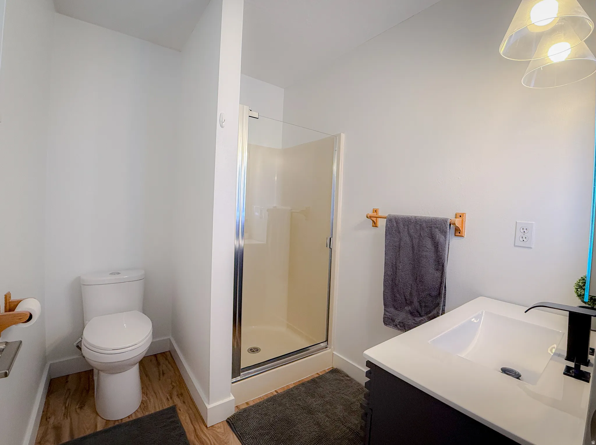 Bathroom featuring a glass-enclosed shower, white vanity with integrated sink, black hardware, wood-finish flooring, and bright pendant lighting
