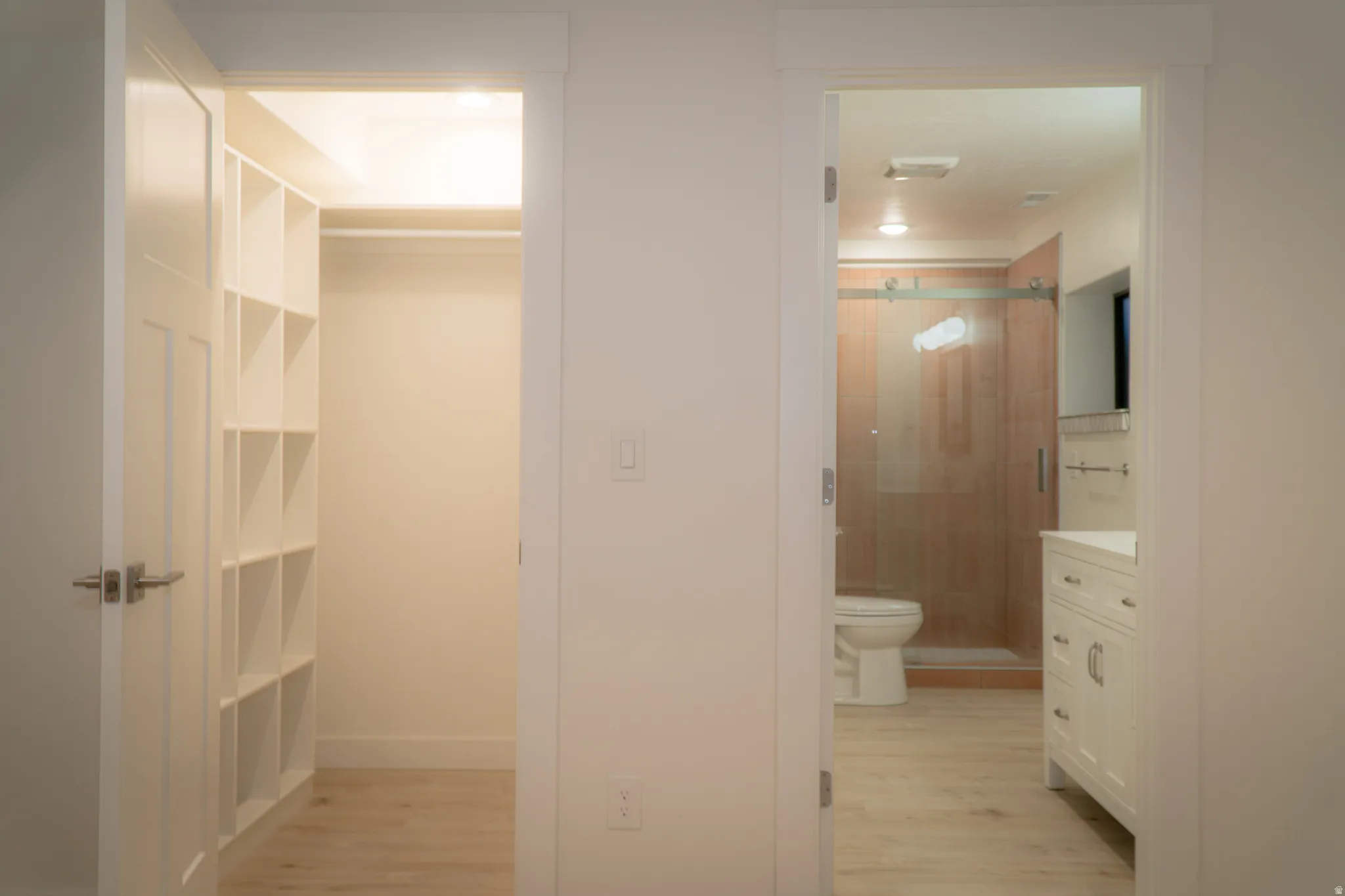Walk-in closet with built-in shelving, wood-finish flooring, and a nearby bathroom featuring a glass-enclosed shower with wood-tone tile, a white vanity with storage, and a toilet
