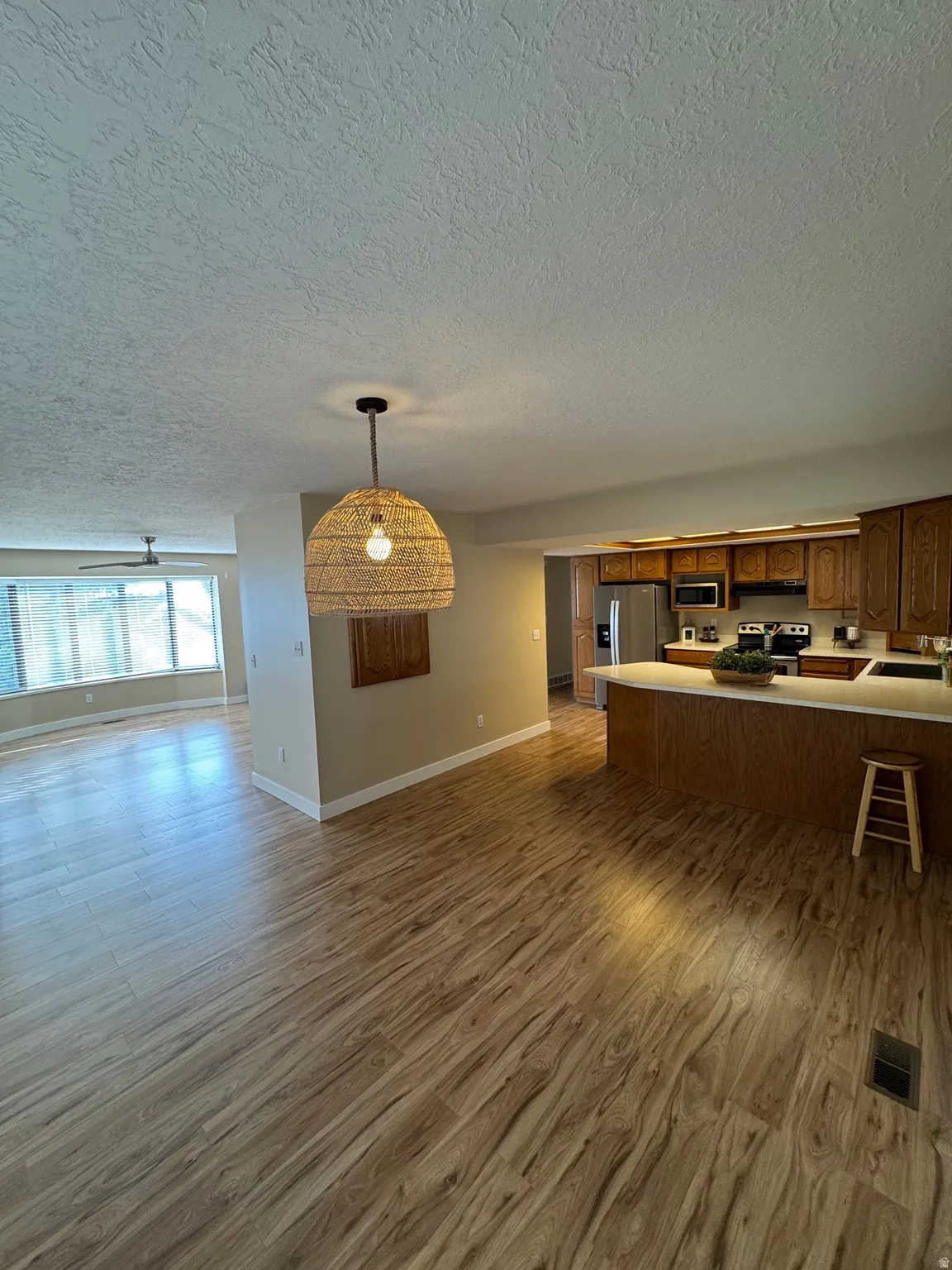 Spacious open-concept living area featuring continuous wood-finish flooring, a modern woven pendant light fixture, and a kitchen with wood cabinetry and stainless steel appliances
